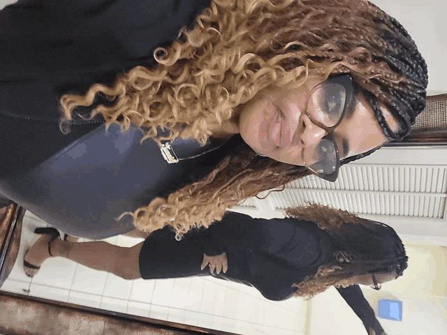 a woman with curly hair and glasses looks at her reflection in the mirror