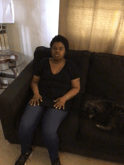 a woman sits on a black couch with a cat laying on the couch
