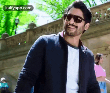 a man wearing sunglasses and a jacket is smiling while standing in front of a building .