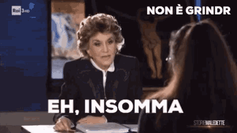 a woman is sitting at a desk talking to another woman with the words eh insomma written on the screen
