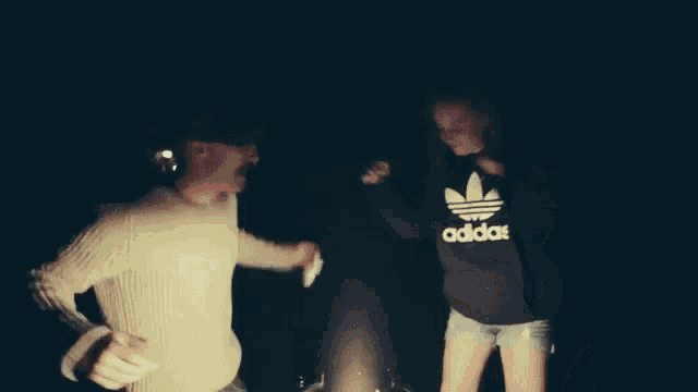 a man and a woman are dancing in front of a fire and the woman is wearing an adidas hoodie