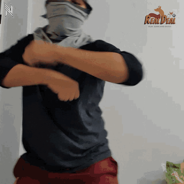 a man wearing a scarf around his face is standing in front of a wall that says real deal