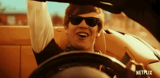 a man wearing sunglasses is driving a car with the word netflix on the steering wheel