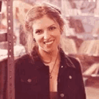 a woman in a black jacket is smiling for the camera while standing next to a shelf .