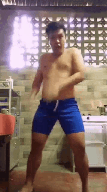 a shirtless man in blue shorts is dancing in a kitchen in front of a window .