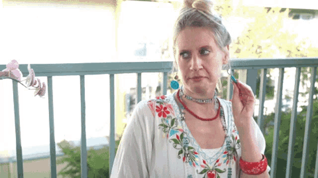 a woman wearing a necklace and earrings is standing on a balcony with flowers in the background