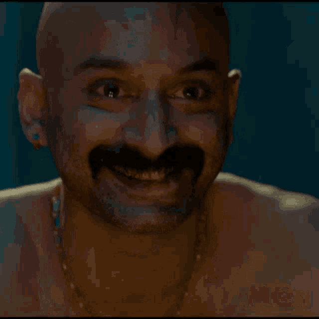 a shirtless man with a mustache and earrings smiles