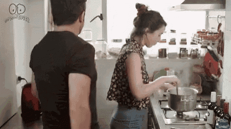 a man and a woman are cooking in a kitchen . the woman is stirring a pot while the man looks on .