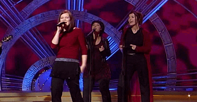 a group of women singing on a stage with a clock behind them
