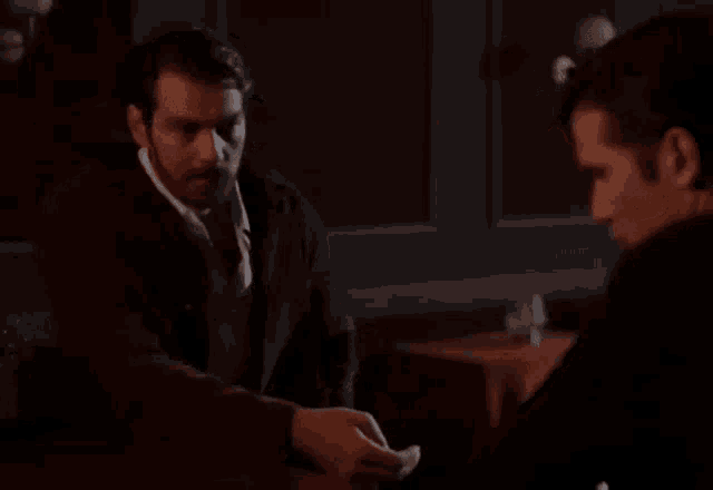 two men are sitting at a table in a dark room and one of them is smoking a cigarette