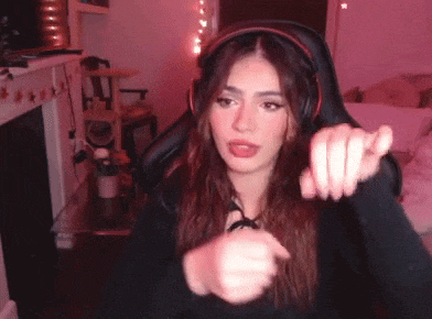 a woman wearing headphones is sitting in a gaming chair and pointing at the camera .