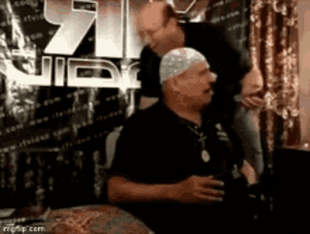two men are standing next to each other in a room . one of the men is wearing a hat .