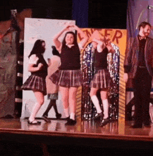 a group of girls in school uniforms are dancing on a stage in front of a man in a suit .