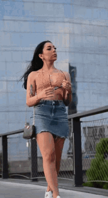 a woman wearing a denim skirt and a crop top is walking on a bridge
