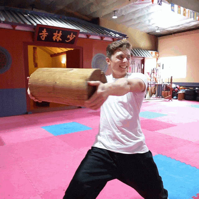 a man in a white shirt is holding a large wooden cylinder in front of a building with chinese writing on it