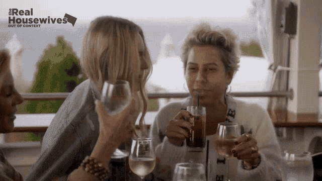 a group of women are sitting at a table drinking wine from glasses with a real housewives logo behind them