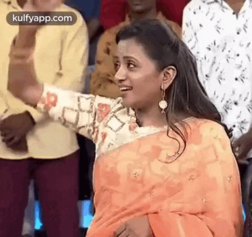 a woman in an orange saree is standing in front of a crowd of people and smiling .