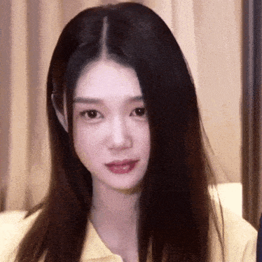 a woman with long black hair is wearing a yellow shirt and looking at the camera