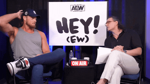 two men sitting in front of a sign that says hey