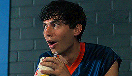 a young man is drinking through a straw from a cup while sitting in front of a brick wall .