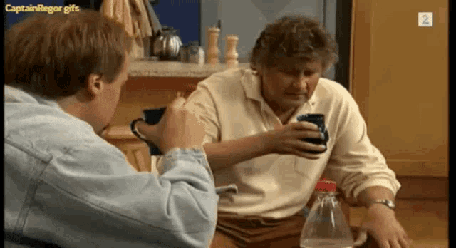 two men are sitting at a table with a bottle of water and a cup of coffee with the number 2 on the bottom right corner
