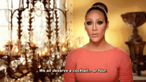 a woman in a pink dress is standing in front of a chandelier and saying we all deserve a cocktail or four .