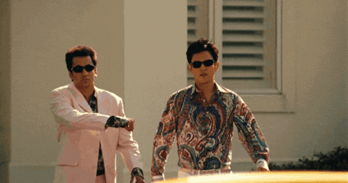 two men wearing sunglasses and paisley shirts stand in front of a building
