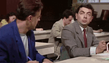 a man in a suit and tie is sitting at a desk in a classroom with another man .