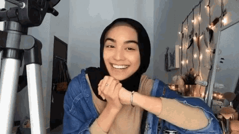 a woman wearing a hijab and a denim jacket is smiling in a room .