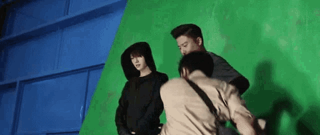 three men are standing in front of a green screen . one of the men is wearing a hooded jacket .