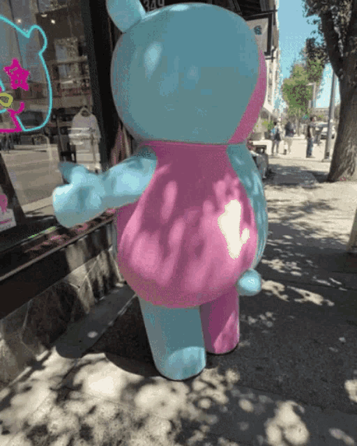 a blue and pink teddy bear is standing on a sidewalk in front of a store
