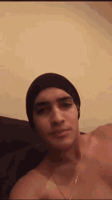 a shirtless man wearing a black beanie and a gold chain looks at the camera