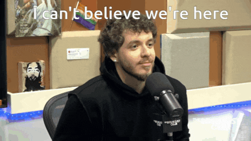 a man sitting in front of a microphone with the words " i can 't believe we 're here " behind him