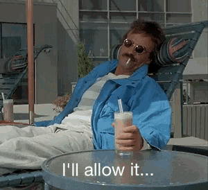 a man in a blue jacket sits in a chair holding a drink and a cigarette