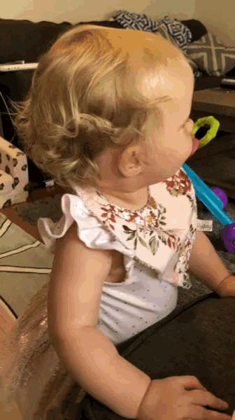 a baby girl wearing a white floral top is sitting on the floor