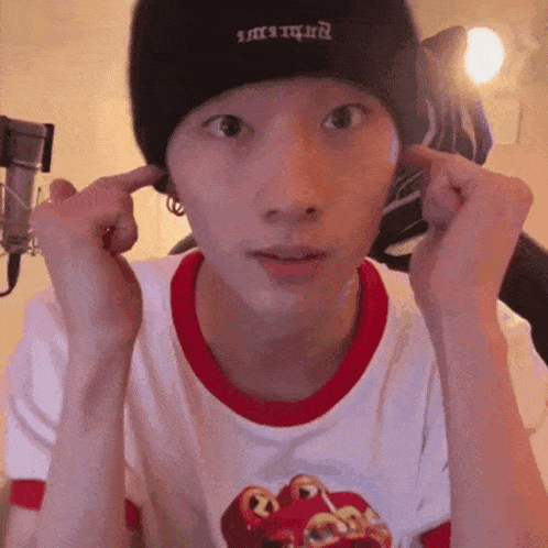 a young man wearing a black beanie and a white shirt is covering his ears with his hands .
