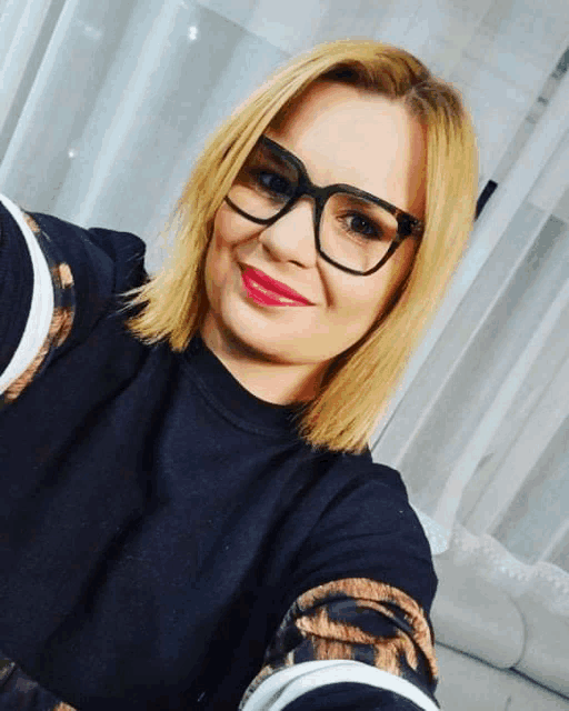 a woman wearing glasses takes a selfie in front of a white curtain