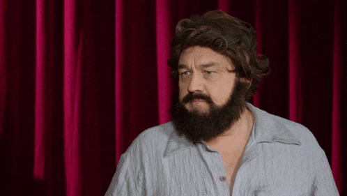 a man with a beard is standing in front of a red curtain and says " achso "