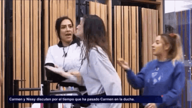 three women are standing in front of a wooden wall and one of them is wearing a blue sweatshirt