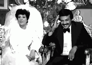 a black and white photo of a bride and groom sitting next to each other on a chair .