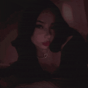 a woman wearing a necklace with a heart shaped pendant is looking at the camera in the dark .