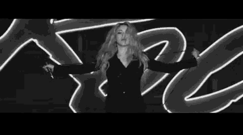 a black and white photo of a woman with long blonde hair standing in front of a neon light .