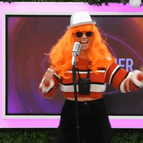 a woman in an orange wig is singing into a microphone in front of a screen that says " er "
