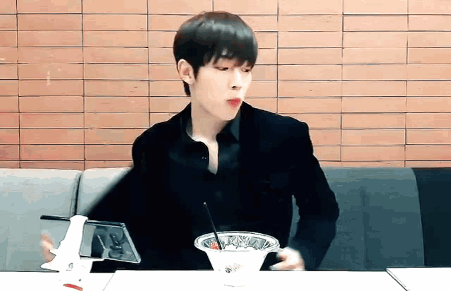 a young man is sitting at a table with a bowl of food and a phone .