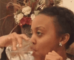 a woman is drinking water from a glass while sitting at a table with a vase of flowers in the background .