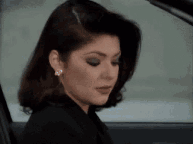a woman in a black jacket and earrings is sitting in a car looking out the window .