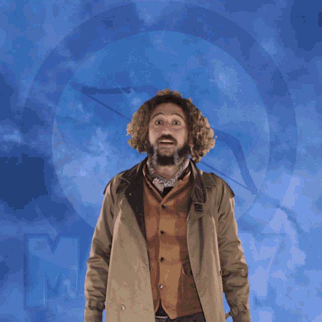 a man with a beard wearing a trench coat stands in front of a blue sky with the letter m visible