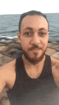 a man with a beard wearing a black tank top is standing in front of the ocean