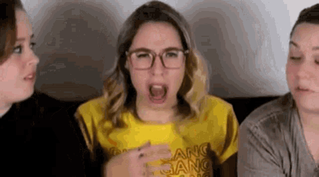 three women are sitting on a couch and one of them is wearing glasses and a yellow t-shirt .