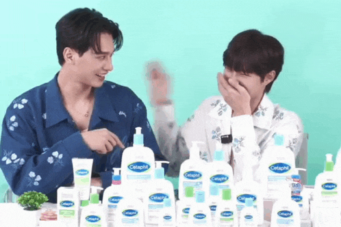 two young men are sitting at a table with bottles of cetaphil products on it .